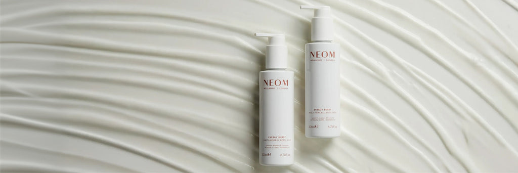NEOM Energy Burst Multi-Mineral Body Milk bottles on textured cream background.