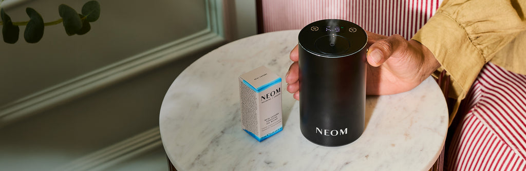 Hand holding NEOM Pod Mini+ portable essential oil diffuser on marble table with Real Luxury essential oil blend.