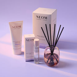 NEOM bath and body products including a body butter, sleep mist, reed diffuser, and packaging on a light purple background