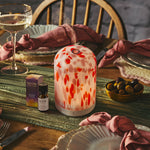 Decorative clementine pod diffuser on a table with wine glasses, olives, and a bottle of essential oil.