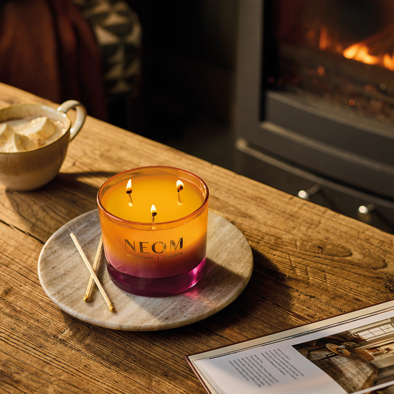 NEOM Cosy Nights 3 Wick Candle on a wooden table with a fireplace in the background