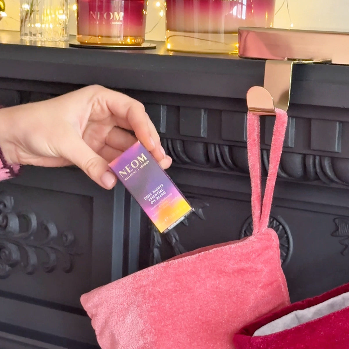 Person placing NEOM Cosy Nights Essential Oil Blend box into pink Christmas stocking on decorated mantel.