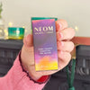 Hand holding NEOM Cosy Nights Essential Oil Blend box with pink and purple design in festive setting