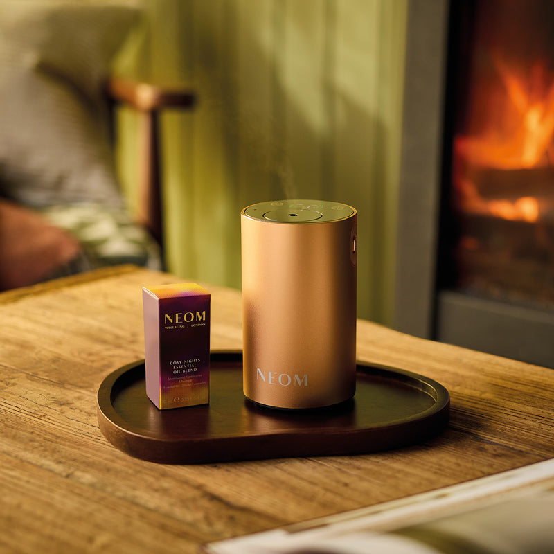 NEOM Pod Mini+ and Cosy Night's Essential Oil Blend on a wooden table with a fireplace in the background