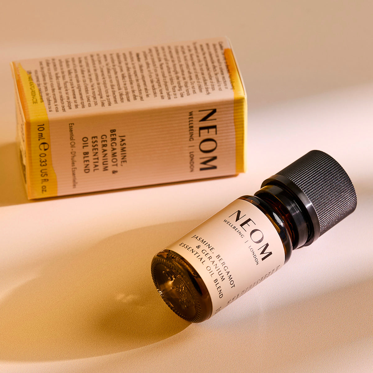 NEOM Jasmine, Bergamot & Geranium essential oil blend bottle lying on a surface with its product box behind it.