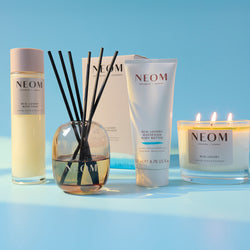 NEOM product range including bath foam, reed diffuser, body butter, and candle on a blue background