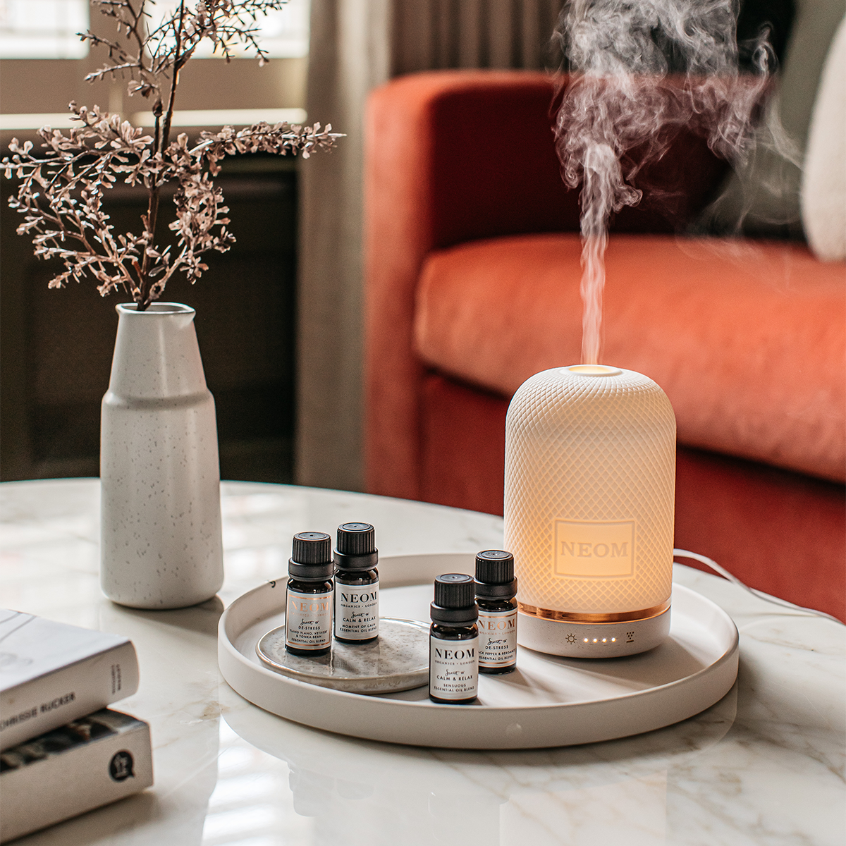 Neom essential oil diffuser on a marble table with Neom bottles and a vase in the background.