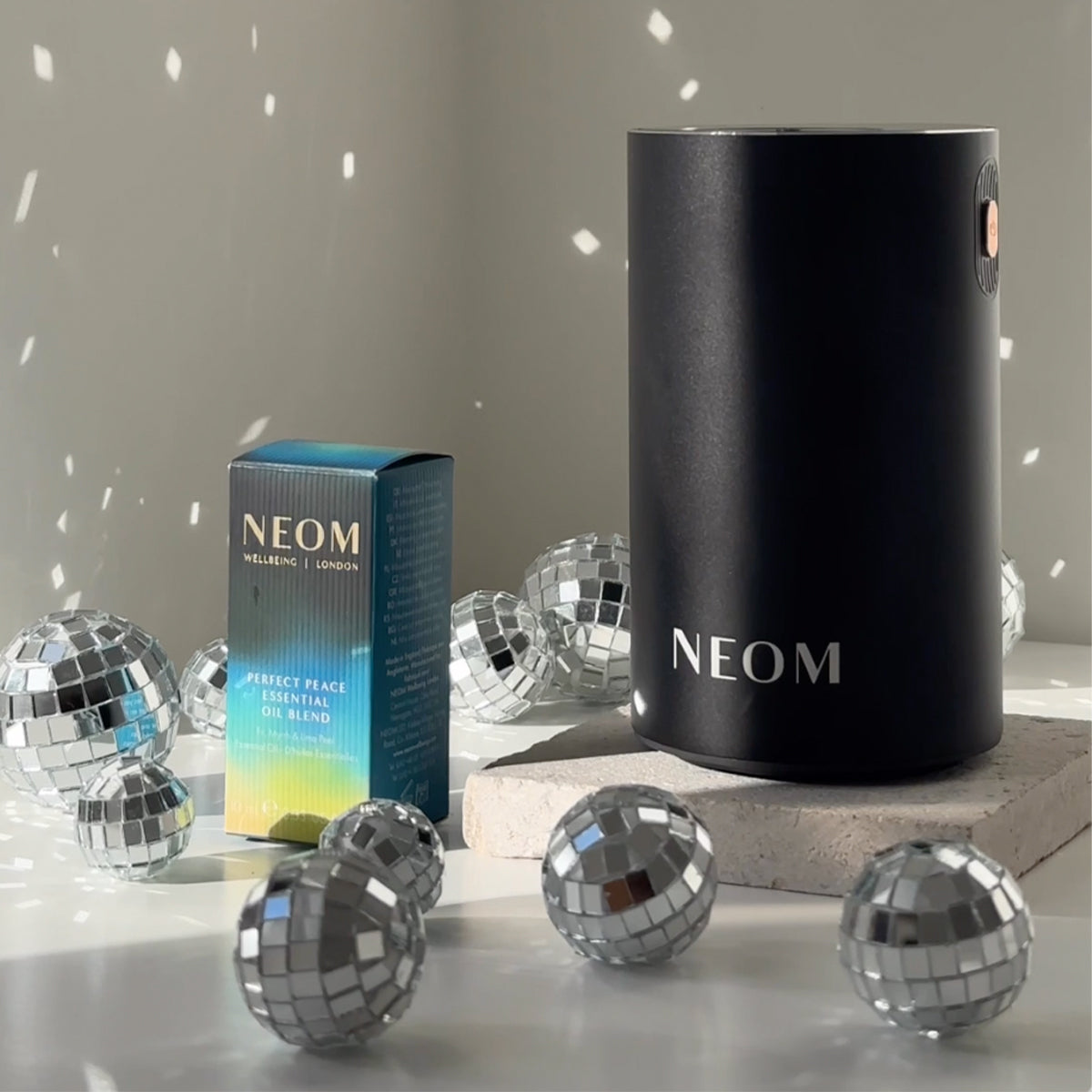 NEOM Perfect Peace Essential Oil Blend and black Wellbeing Pod Mini surrounded by sparkling disco balls.