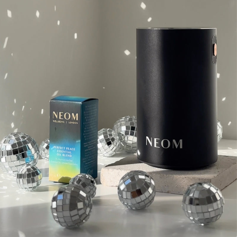NEOM Perfect Peace Essential Oil Blend and black Wellbeing Pod Mini surrounded by sparkling disco balls.