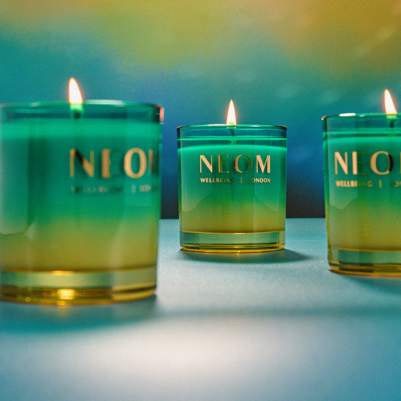Three lit candles in green glass jars with 'NLCM' branding on a blurred background