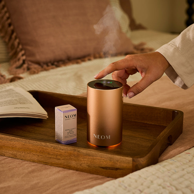 NEOM diffuser on a wooden tray with a hand interacting, surrounded by a cozy indoor setting.