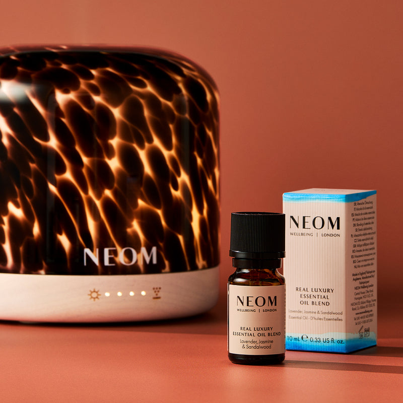 NEOM diffuser, essential oil blend bottle, and packaging on a warm brown background